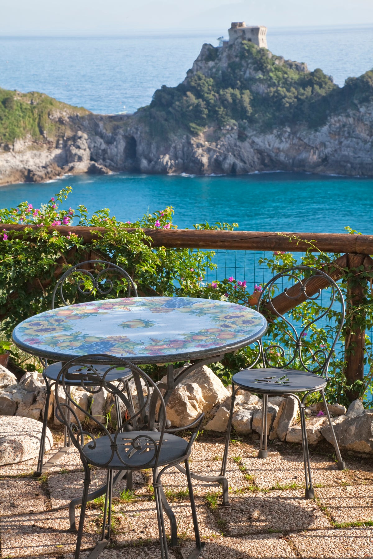 Amalfi Coast: a hand-painted ceramic table and iron chairs on a terrace above a turquoise cove, flowering vines along the rail.