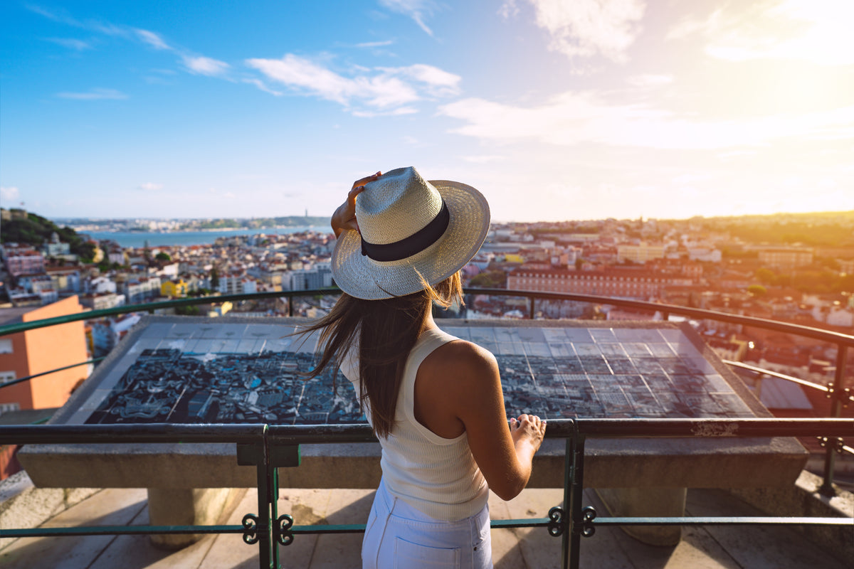 Lisbon: a woman in a straw hat stands at a hilltop viewpoint, looking out over rooftops toward the river.